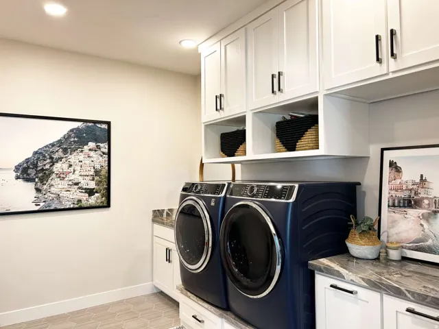 Dedicated laundry room featuring side-by-side front-load washers and dryers in navy blue, white shaker-style cabinetry with brushed black hardware, open shelving with woven storage baskets, quartz countertop workspace, recessed lighting, and framed artwor
