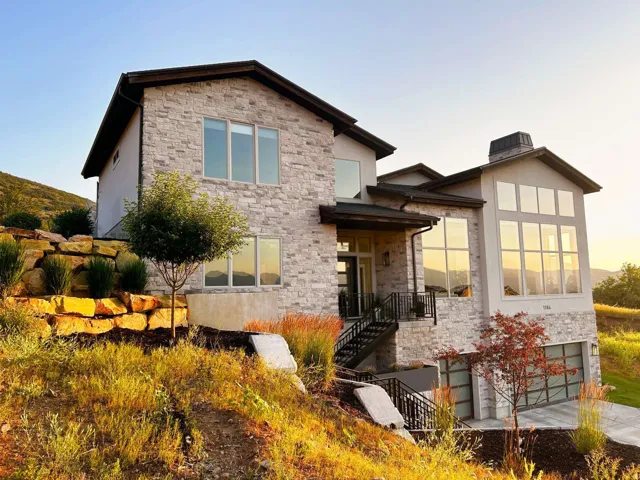 Contemporary residence featuring a stone and stucco exterior, multiple large windows, a multi-paneled garage door, and a tiered rock retaining wall with landscaping