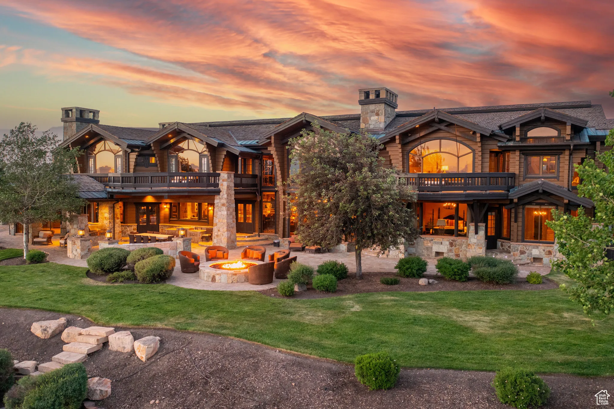 Back of property at dusk with a balcony, a patio, a lawn, stone siding, and a chimney