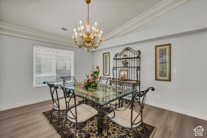 Spacious room featuring a prominent chandelier, light wood-finish flooring, a large window with blinds, crown molding, and neutral wall paint