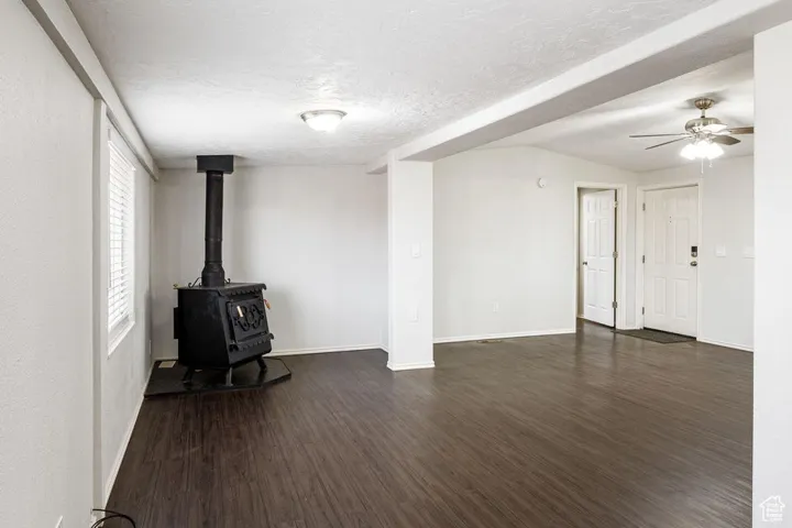 Unfurnished living room with a textured ceiling, dark wood-style flooring, a ceiling fan, baseboards, and a wood stove