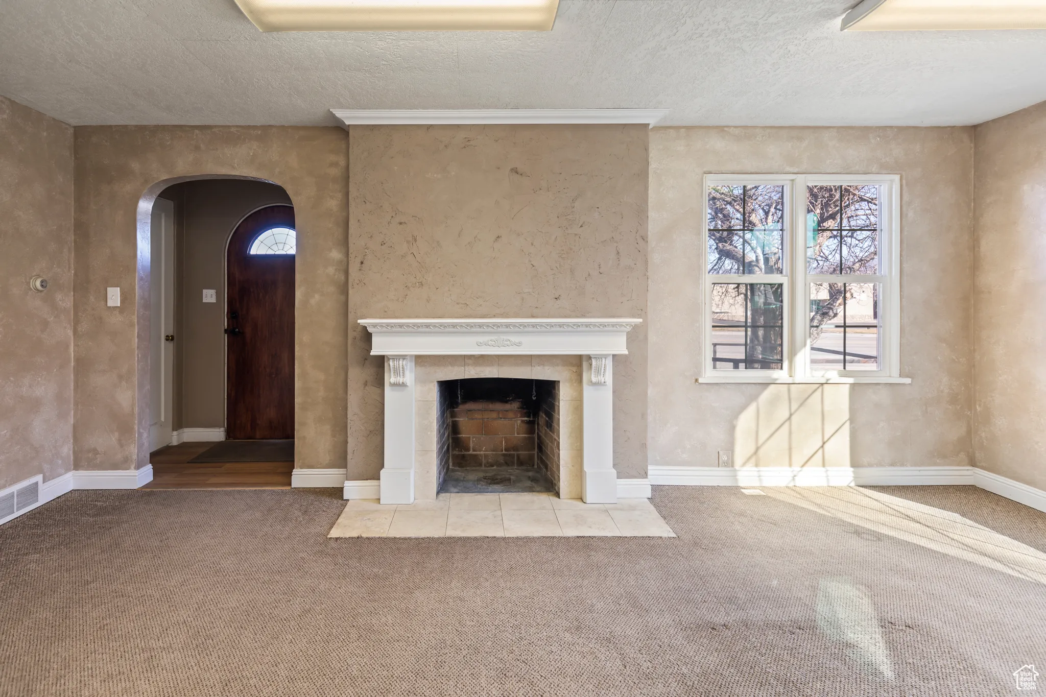 Unfurnished living room featuring a tiled fireplace, a textured ceiling, carpet floors, arched walkways, and baseboards