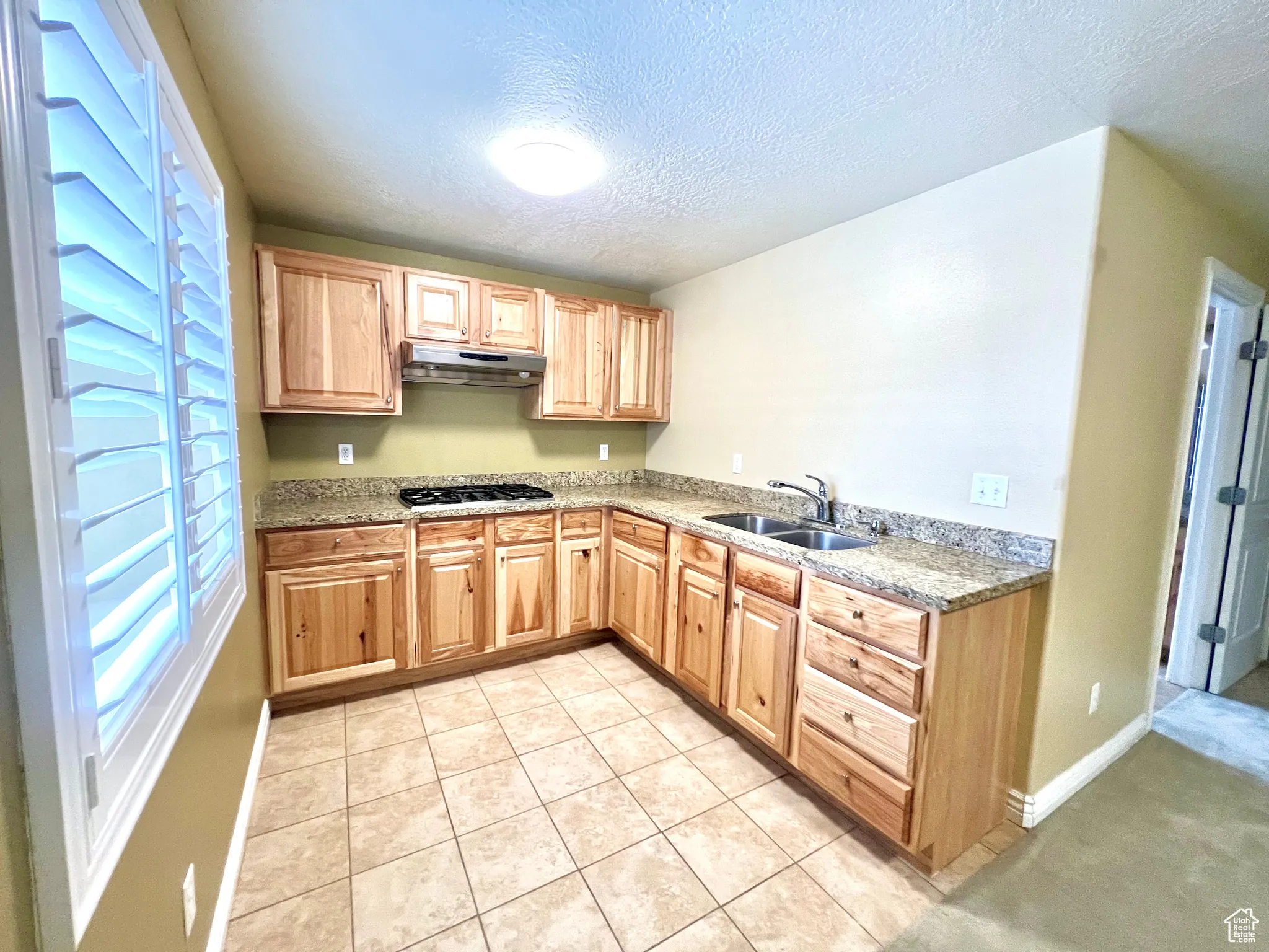 Kitchen featuring light brown cabinetry, under cabinet range hood, a sink, a textured ceiling, and gas stovetop