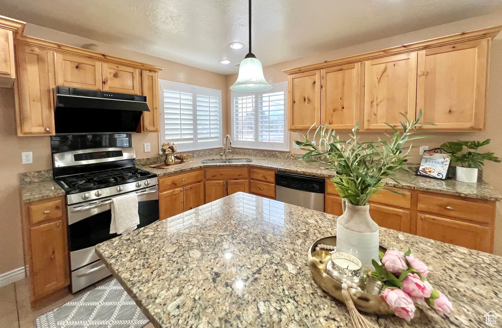 Kitchen with light stone countertops, under cabinet range hood, appliances with stainless steel finishes, hanging light fixtures, and a sink