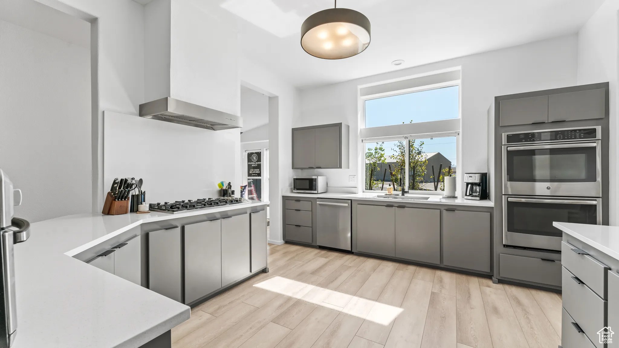 Kitchen featuring gray cabinetry, appliances with stainless steel finishes, wall chimney exhaust hood, light wood finished floors, and light countertops