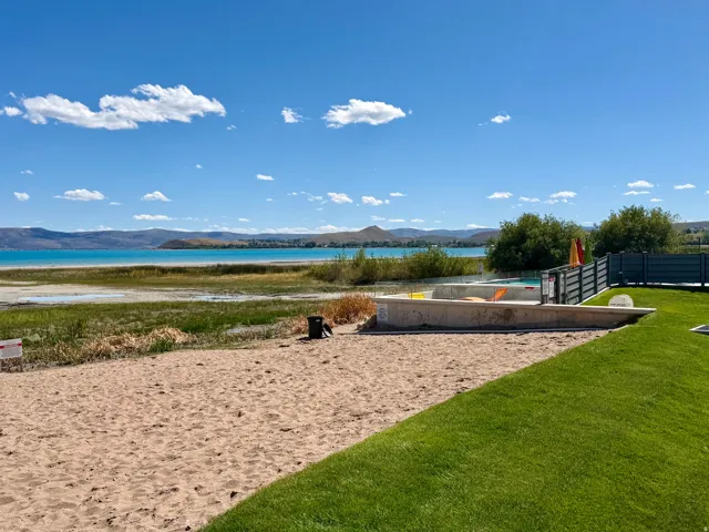Manicured lawn area with a sandy beach section and waterscape beyond, featuring a concrete-edged pool area and dark-toned fencing