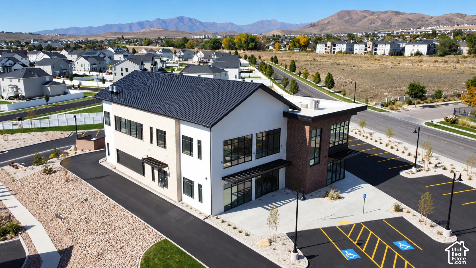 Contemporary multi-story building featuring a white stucco and brick facade, a metal roof, expansive windows, and a dedicated paved parking lot with designated spaces