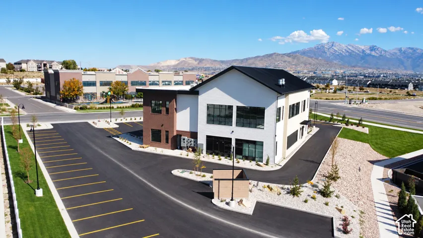 Multi-level commercial structure featuring brick and smooth-panel siding, expansive asphalt parking lot, large windows, and mountain backdrop
