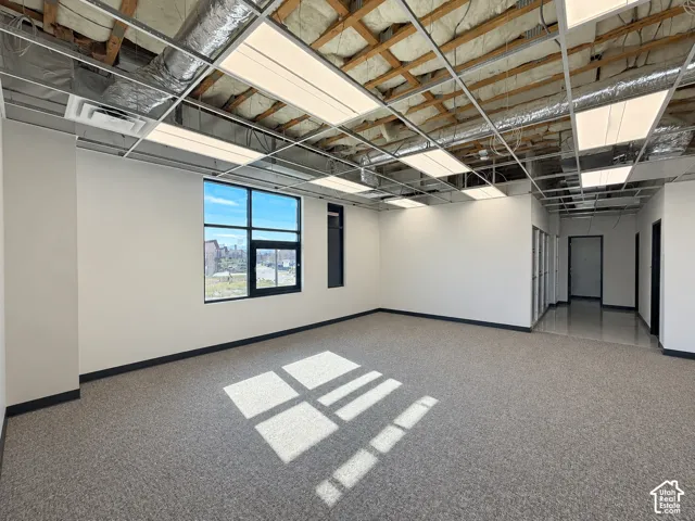Open-concept interior featuring carpeted flooring, large rectangular window, exposed ceiling grid with integrated lighting, and neutral wall finishes