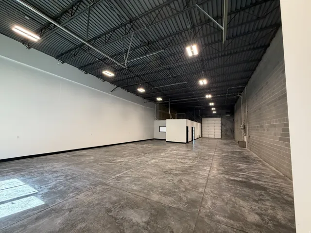 Spacious commercial interior featuring concrete flooring, tall ceilings with exposed metal trusses, bright overhead lighting, a large roll-up door, and a built-in office area