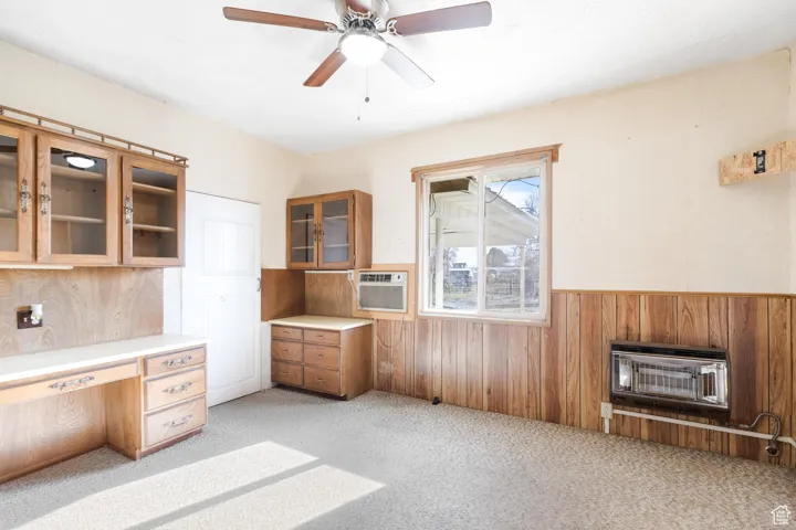 Unfurnished office with wooden walls, heating unit, a ceiling fan, light carpet, and wainscoting