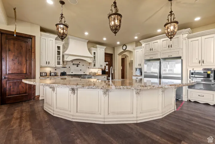 Kitchen featuring dark wood-type flooring, arched walkways, appliances with stainless steel finishes, premium range hood, and backsplash