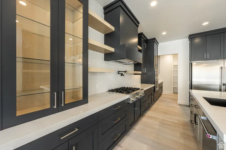 Kitchen featuring appliances with stainless steel finishes, wall chimney exhaust hood, light wood-style flooring, dark cabinetry, and open shelves