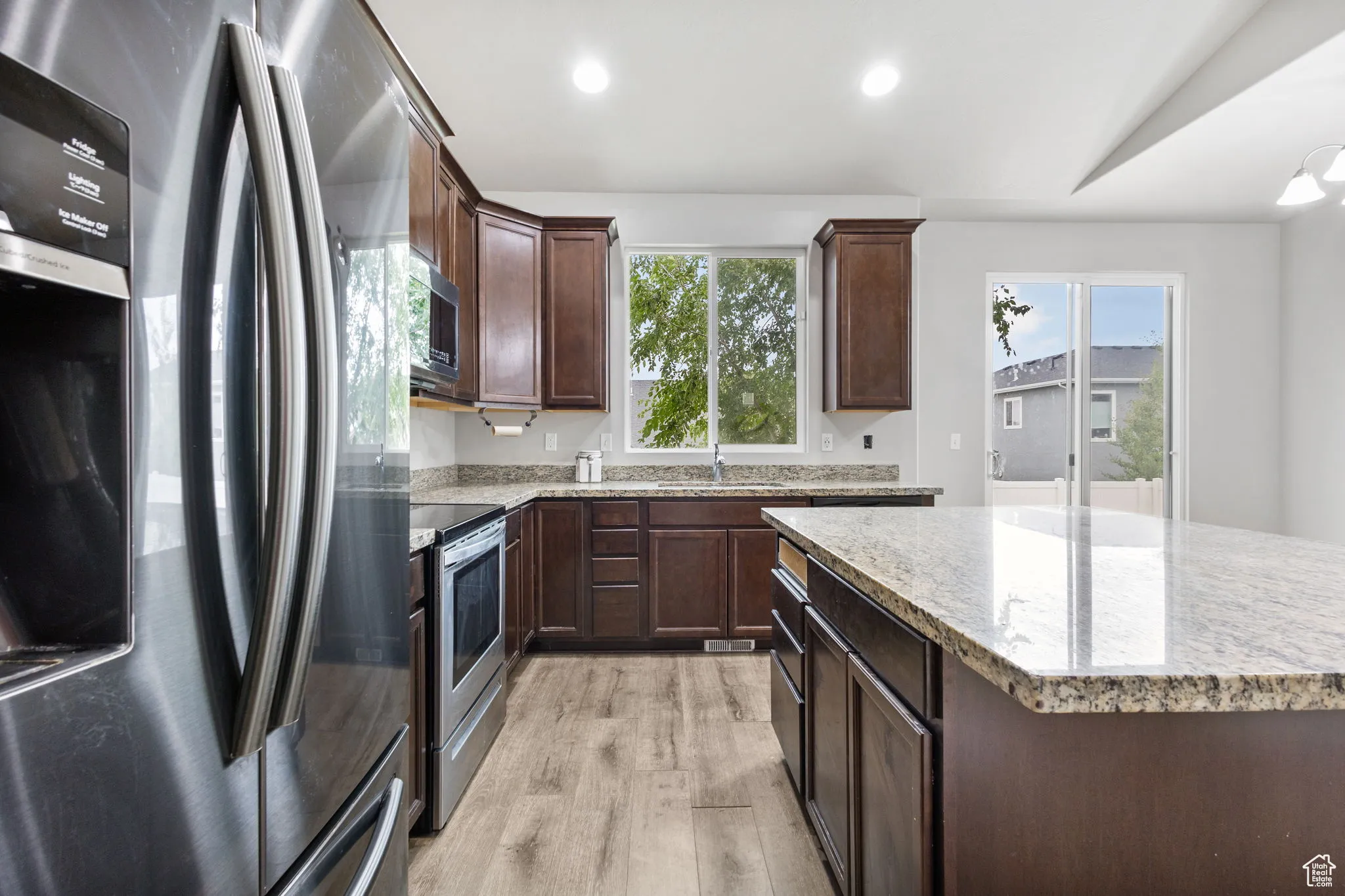 Kitchen featuring appliances with stainless steel finishes, light wood finished floors, dark brown cabinets, a kitchen island, and light stone countertops