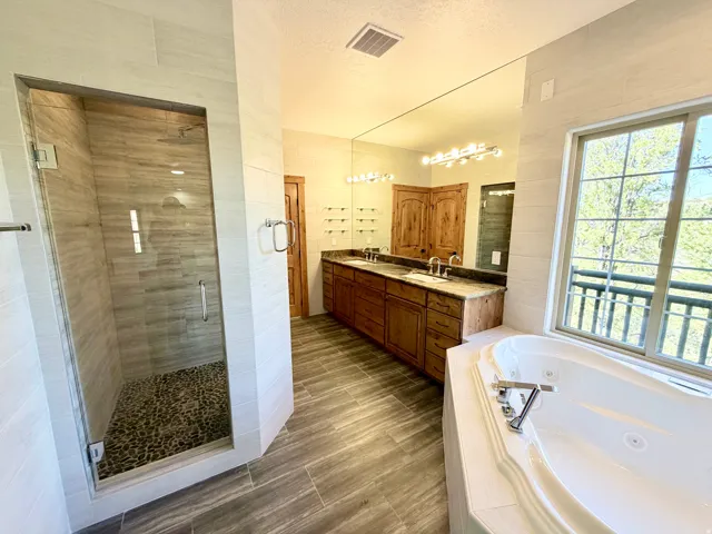Spacious bathroom featuring a glass-enclosed shower with pebble flooring, a large soaking tub, a dual vanity with a stone countertop, a full-wall mirror, and wood-finish flooring