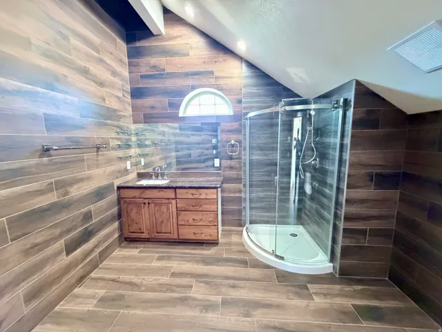 Bathroom featuring wood-finish tile flooring and wall treatment, a curved glass shower enclosure, a wood vanity with a granite-style countertop, and a half-round transom window