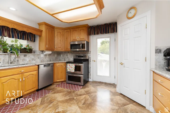 Kitchen with backsplash, light stone countertops, appliances with stainless steel finishes, brown cabinetry, and light stone finish floors