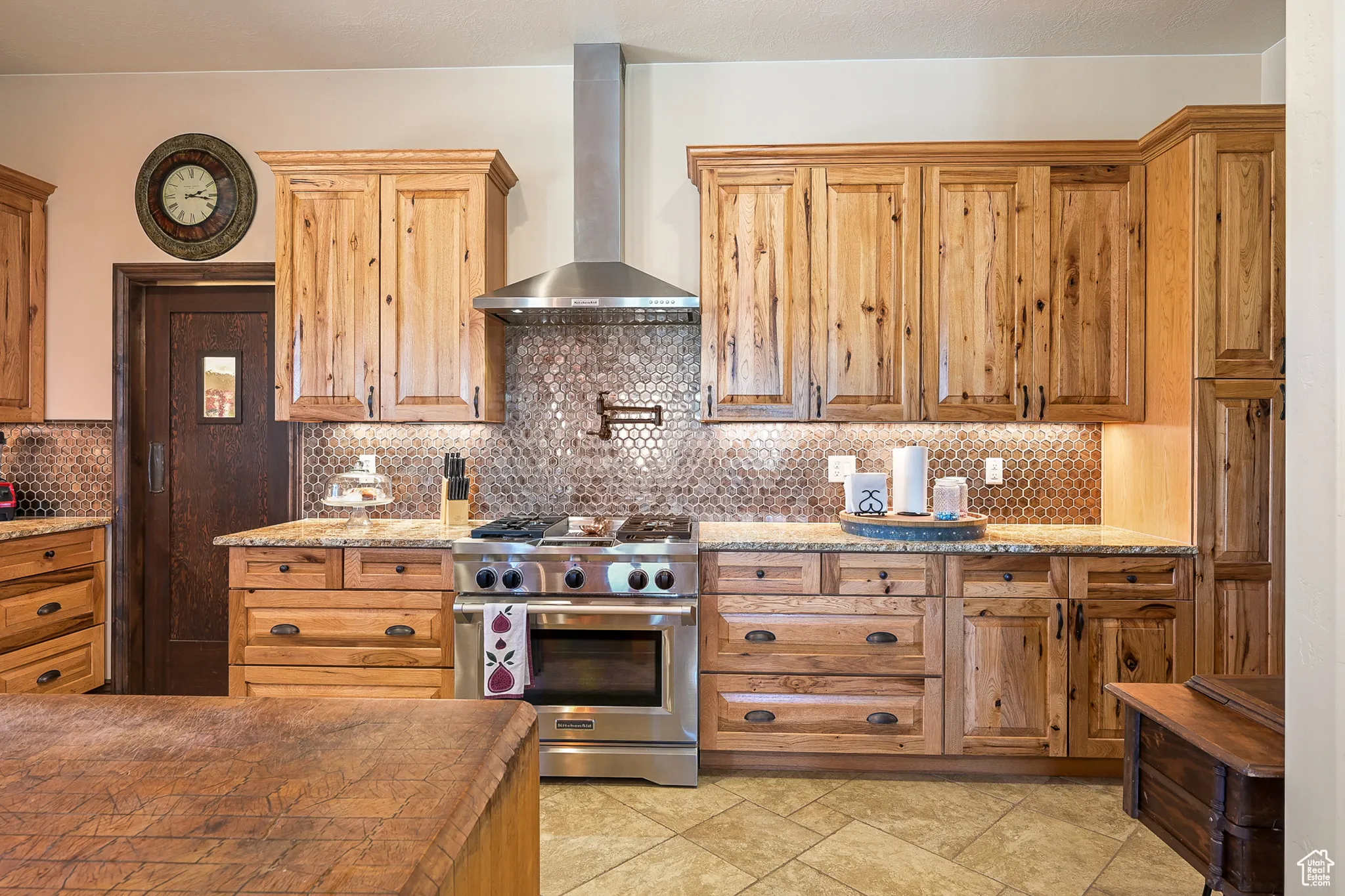 Kitchen with stainless steel range, wall chimney exhaust hood, backsplash, brown cabinets, and light stone countertops