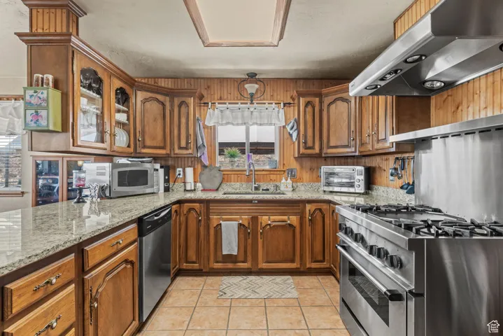 Kitchen featuring range hood, a sink, stainless steel appliances, brown cabinets, and light stone counters