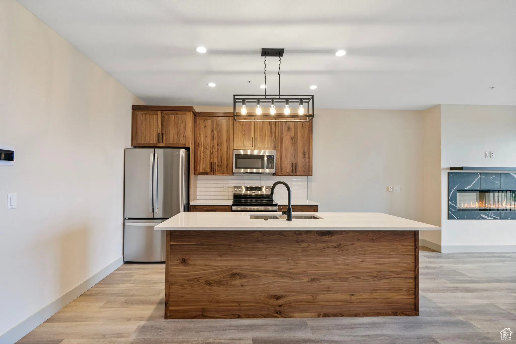 Kitchen with brown cabinetry, tasteful backsplash, appliances with stainless steel finishes, a center island with sink, and hanging light fixtures