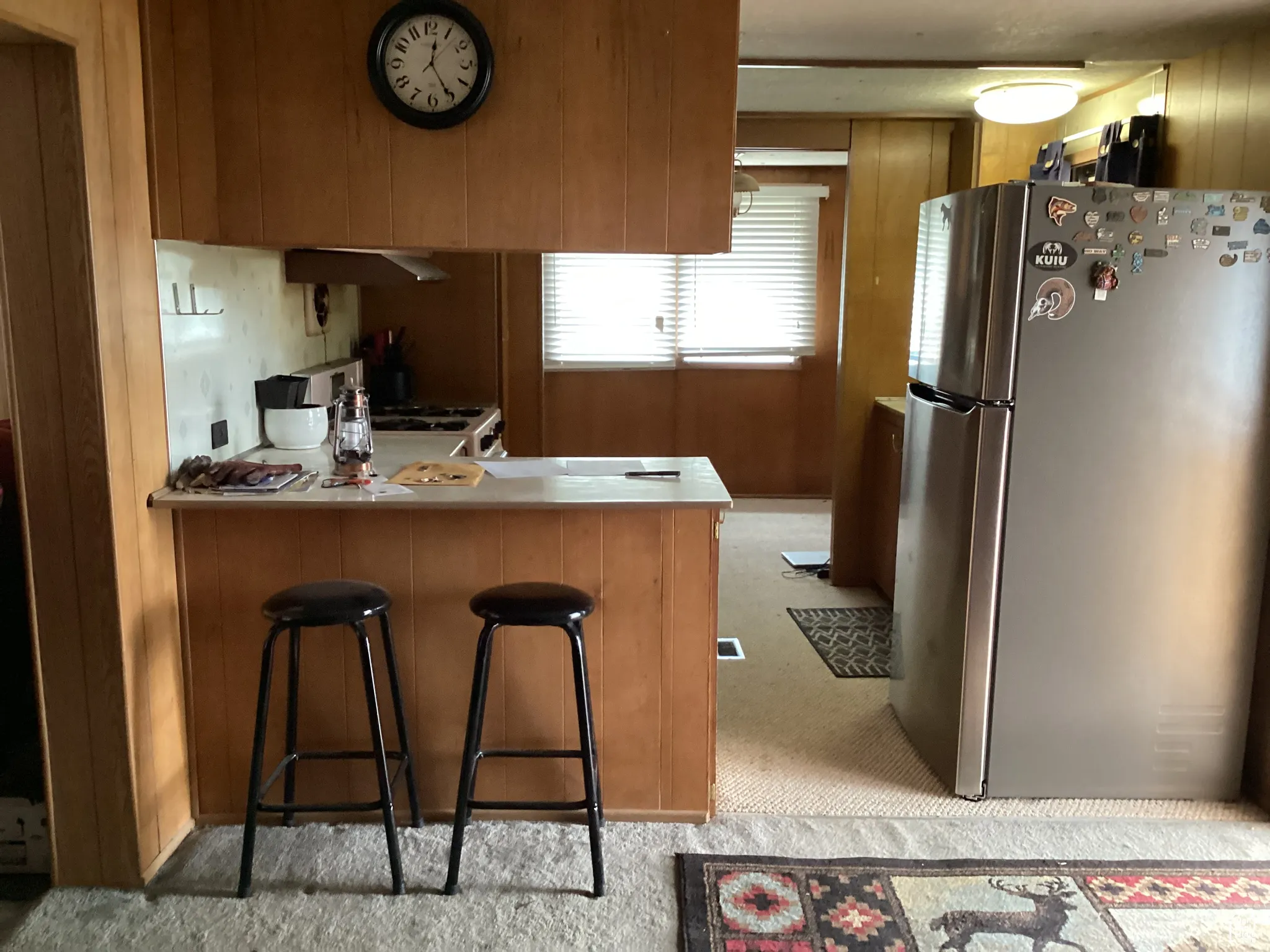 Kitchen featuring freestanding refrigerator, light colored carpet, a peninsula, brown cabinets, and under cabinet range hood