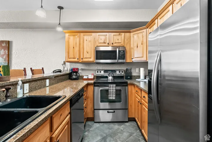 Kitchen with stainless steel appliances, a sink, a textured wall, hanging light fixtures, and dark tile patterned flooring