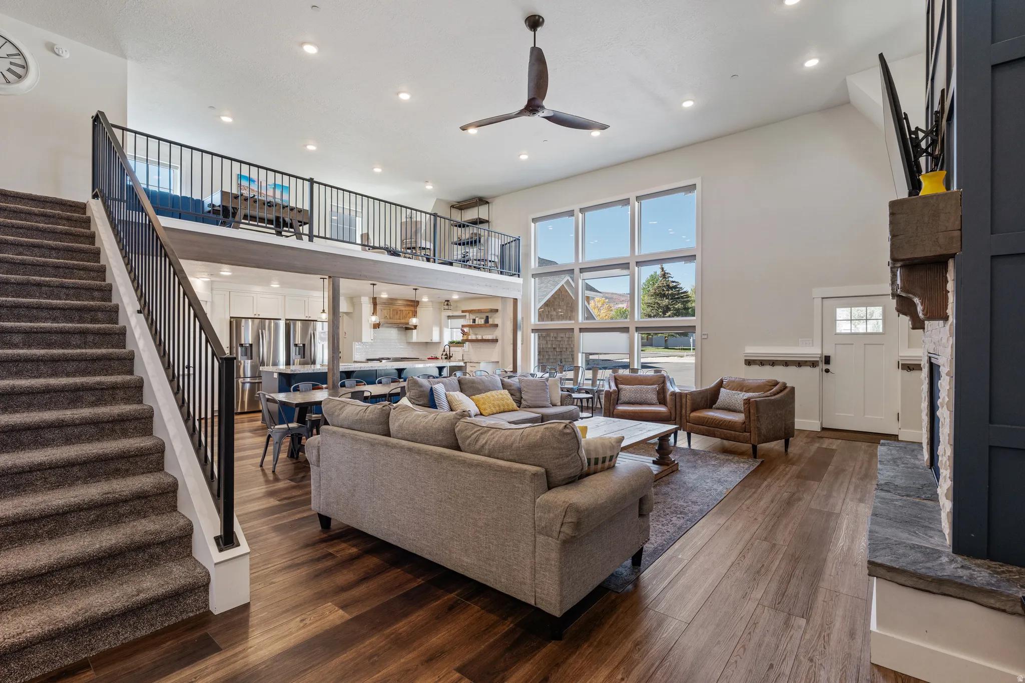 Open-concept living area featuring high ceilings, wood-finish flooring, a stone fireplace with a wood mantle, and a built-in loft space