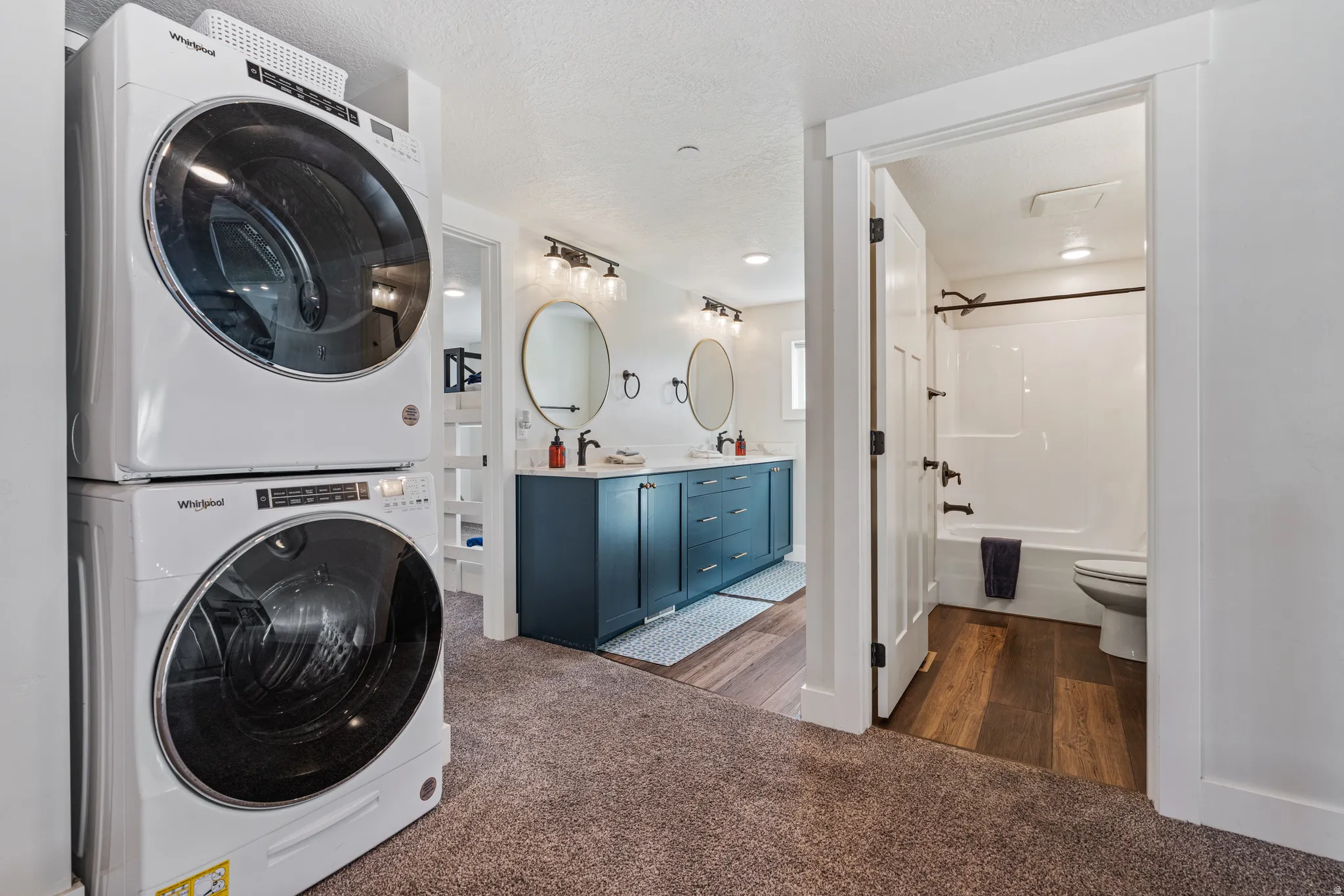 Stackable laundry units, dual vanity with round mirrors, modern wall-mounted light fixtures, tub-shower combination, and wood-finish flooring