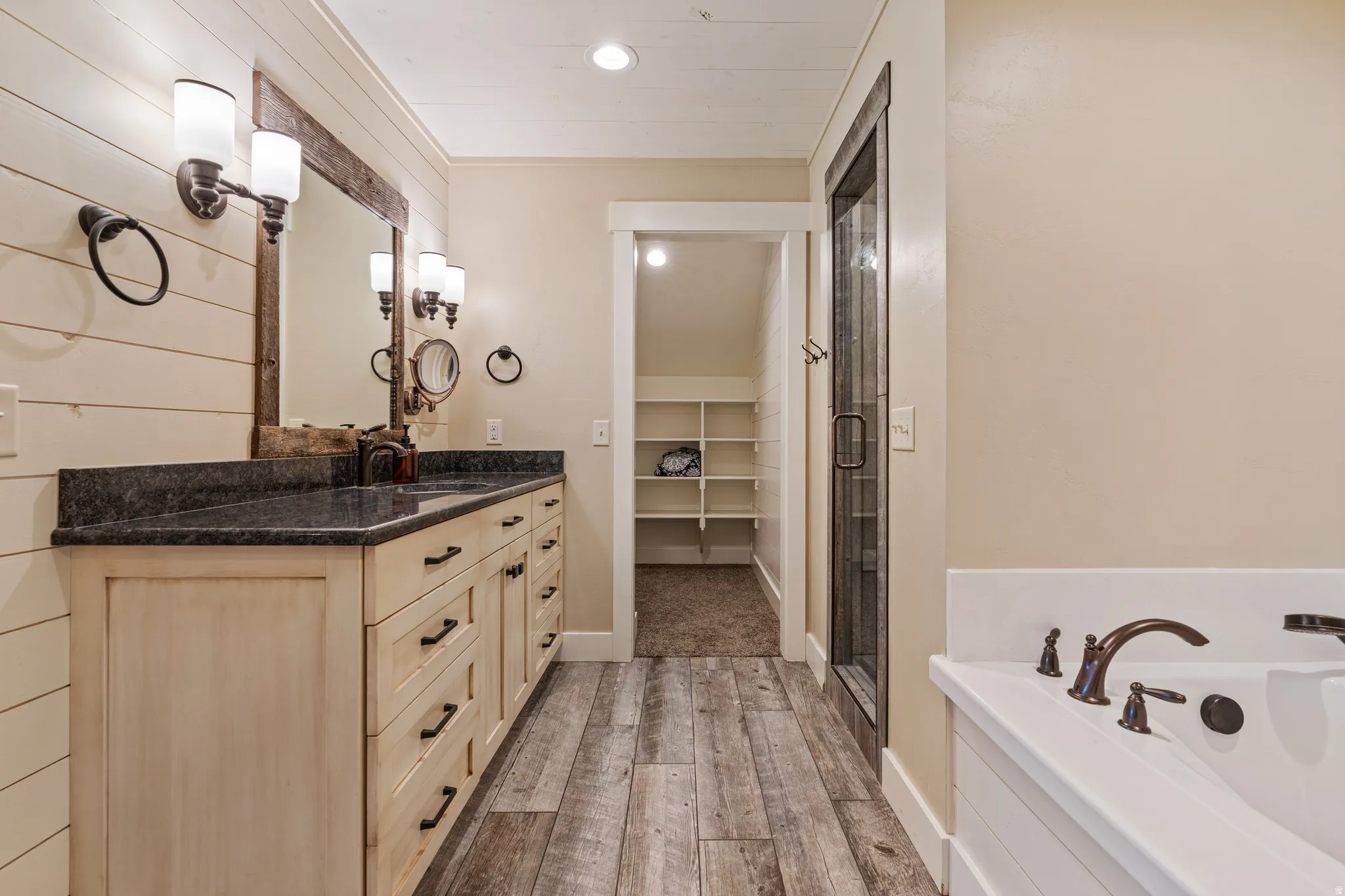 Dual vanity with dark countertop, wood-finish flooring, built-in soaking tub with dark bronze fixtures, and a glass-enclosed shower