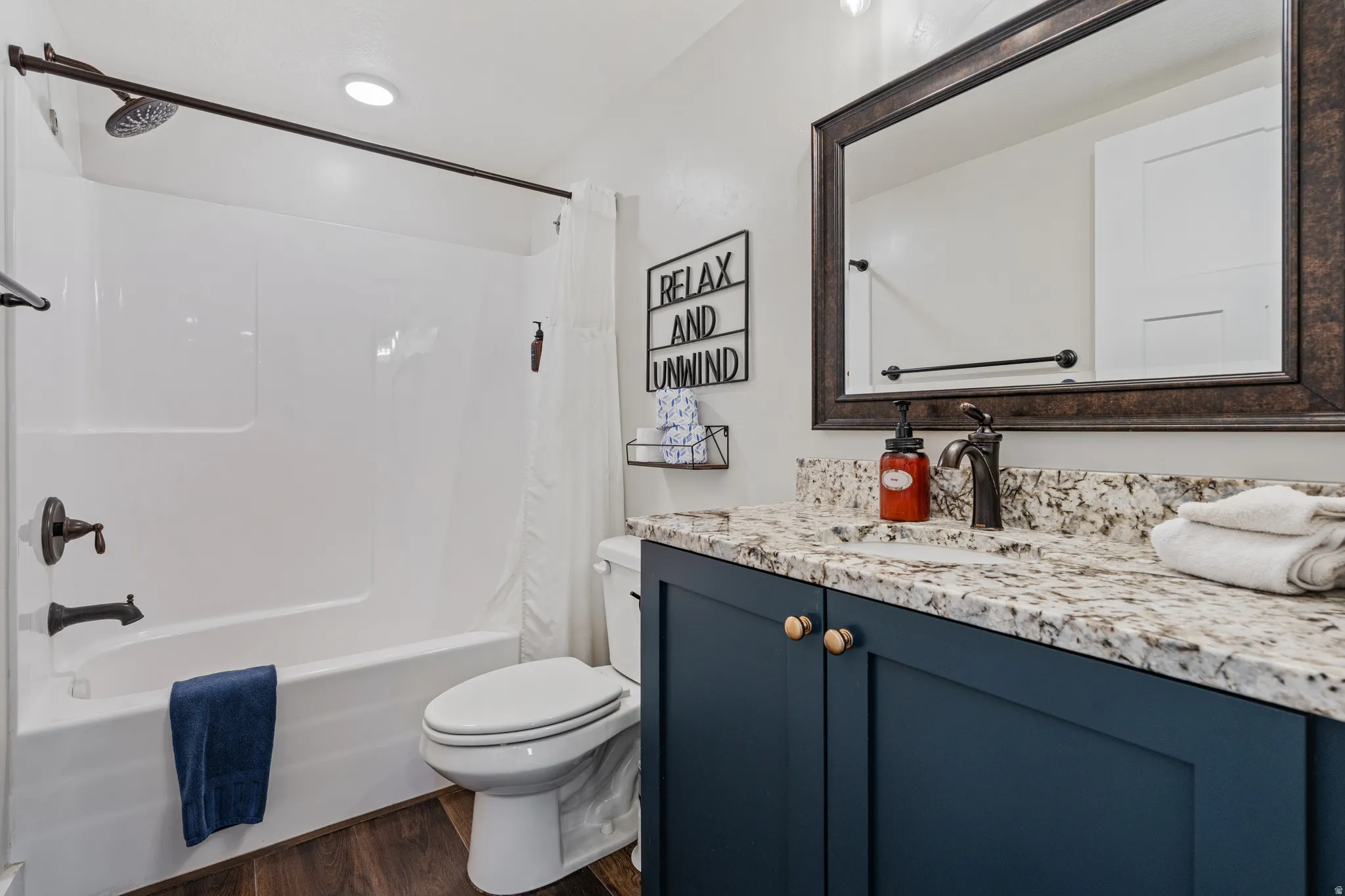 Bathroom featuring a white tub and shower combination with dark bronze fixtures, dark wood-finish flooring, a toilet, a vanity with a light-colored speckled countertop, and a dark blue base cabinet with gold-toned knobs
