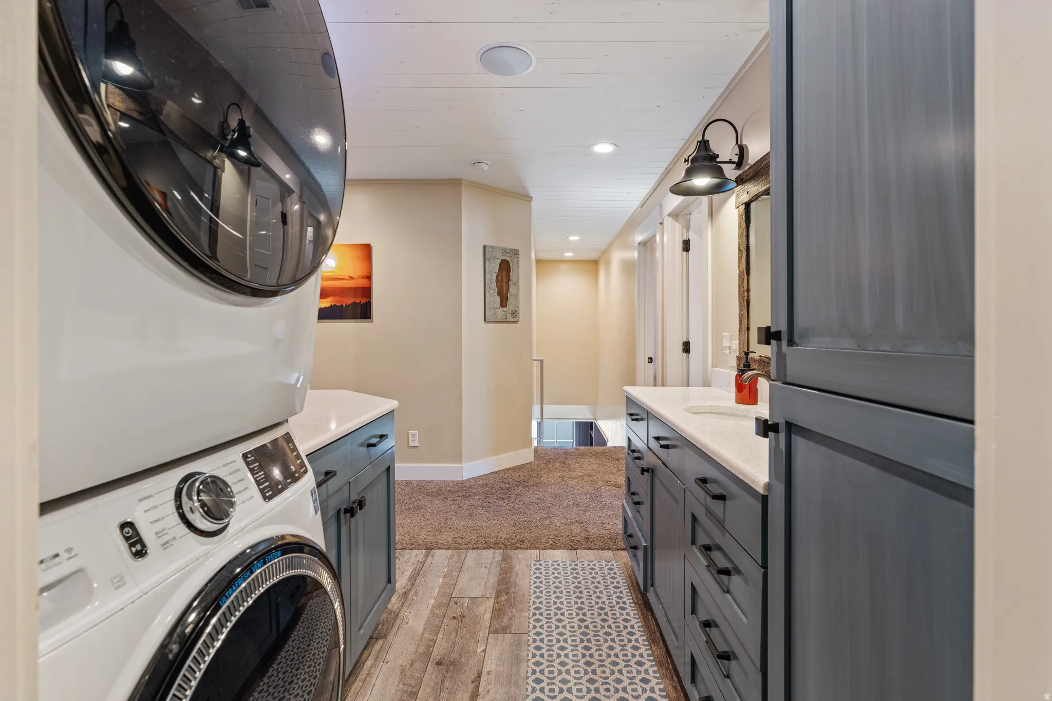 Dedicated laundry room featuring front-load washer and dryer, gray cabinetry with ample storage, wood-finish flooring, decorative geometric runner, recessed lighting, and sconce wall fixtures throughout the galley-style layout