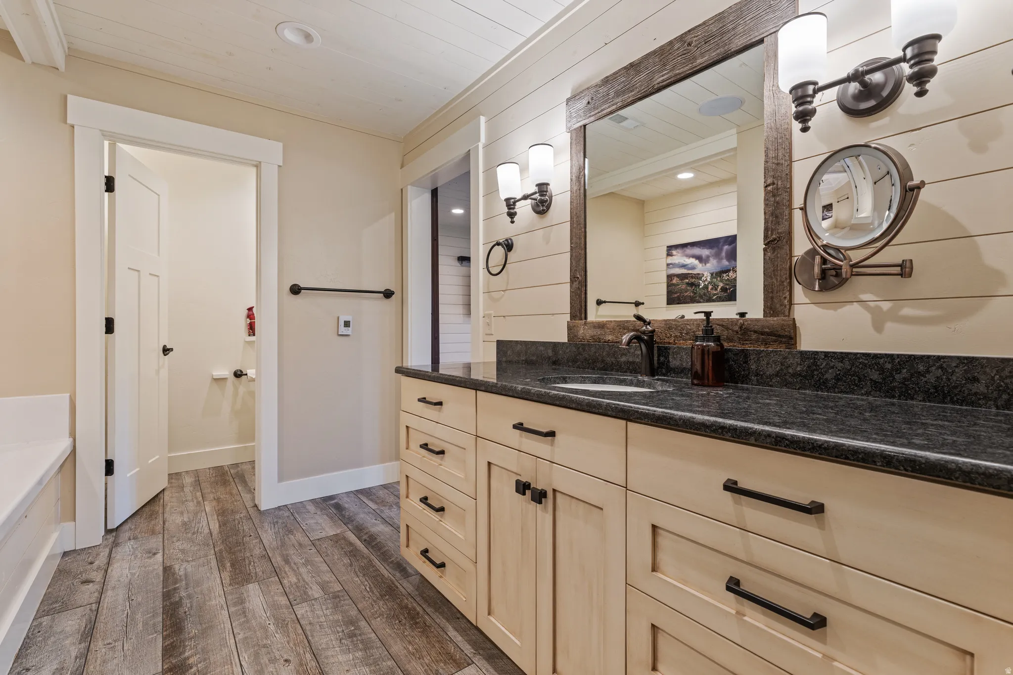 Wood-finish flooring, stone countertop vanity with integrated sink, rectangular mirror with rustic wood frame, and dual wall-mounted light fixtures