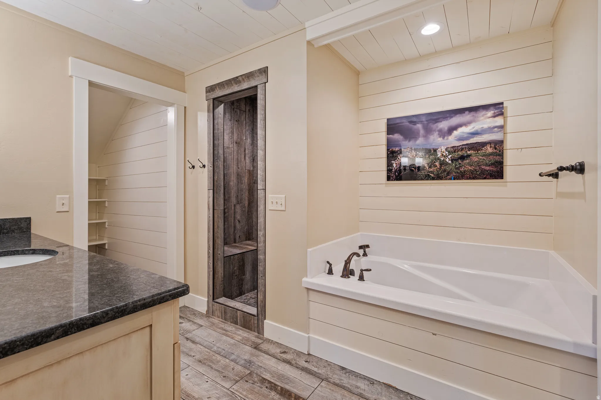 Built-in soaking tub with dark bronze fixtures, wood-finish flooring, single vanity with dark countertop, and built-in shelving