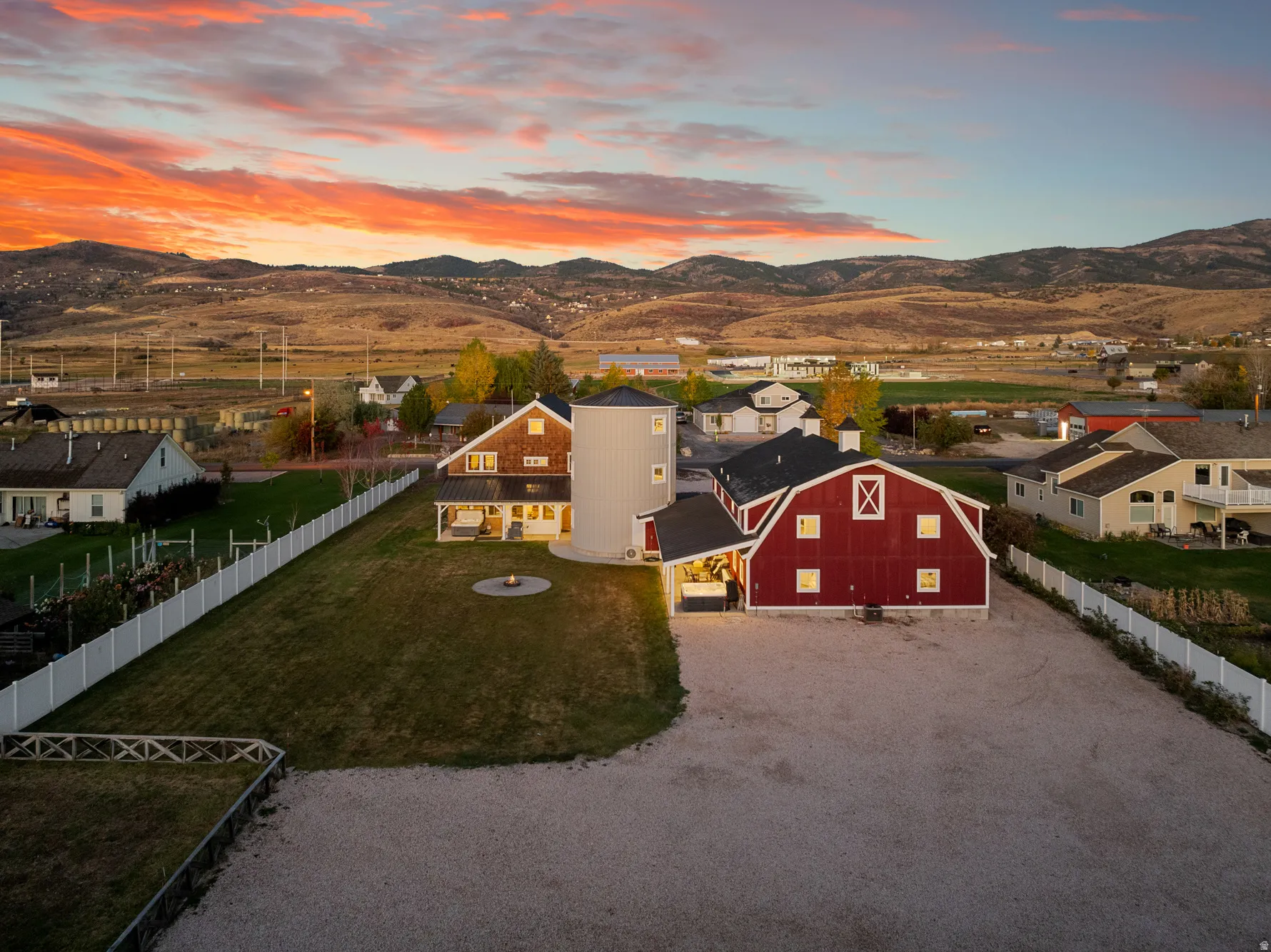 Red barn-style structure, adjacent silo, white picket fencing, gravel driveway, and a backyard fire pit