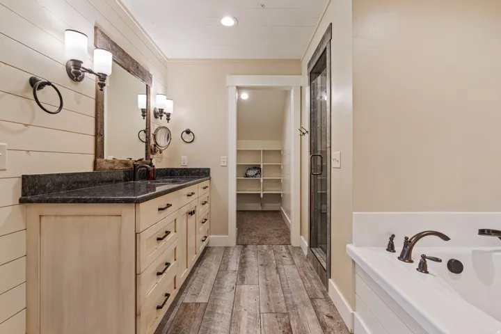 Dual vanity with dark countertop, wood-finish flooring, built-in soaking tub with dark bronze fixtures, and a glass-enclosed shower