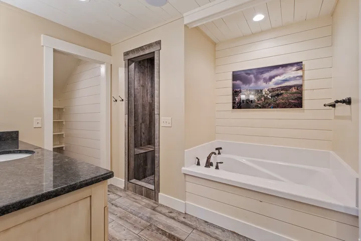 Built-in soaking tub with dark bronze fixtures, wood-finish flooring, single vanity with dark countertop, and built-in shelving