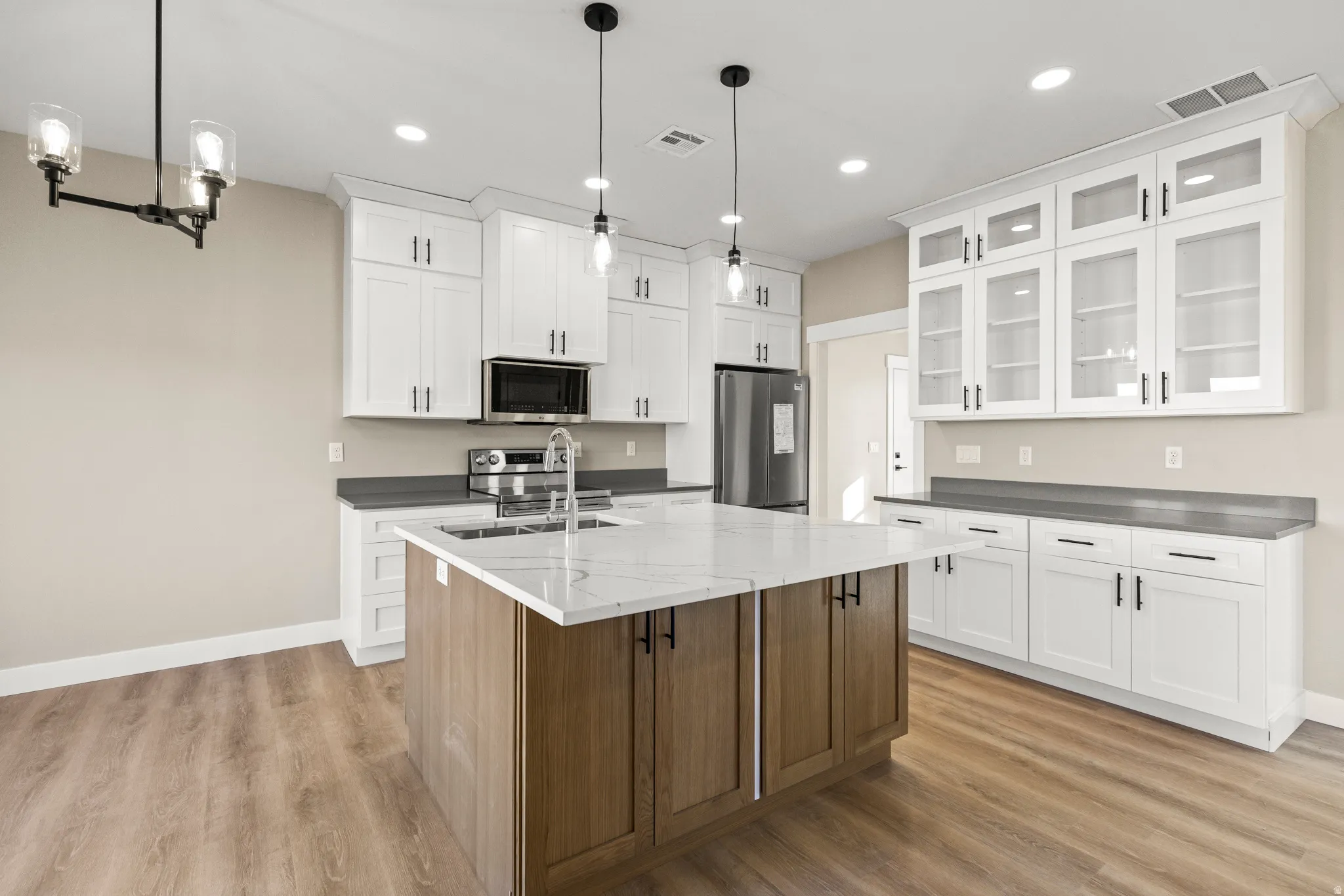 Kitchen with white cabinets, glass insert cabinets, an island with sink, decorative light fixtures, and stainless steel appliances