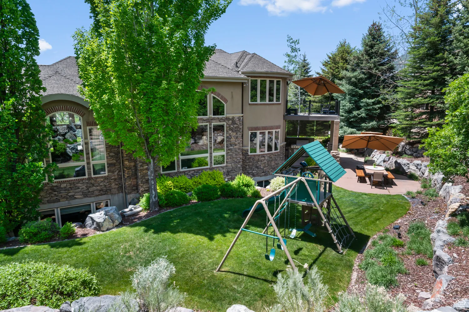 Back of property featuring stone siding, a patio, a playground, a lawn, and a balcony
