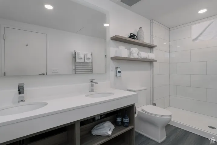 Bathroom featuring toilet, double vanity, a tile shower, wood finished floors, and recessed lighting