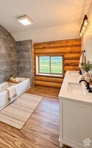 Wood-finish flooring, log wall accents, white double vanity with integrated sinks, and a built-in bathtub with a tiled surround