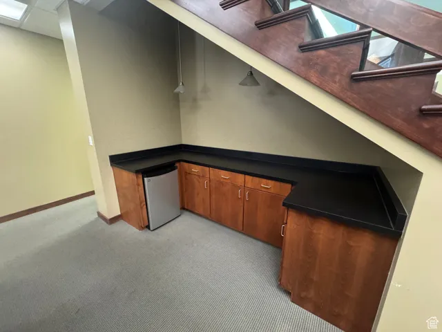 Kitchen with light colored carpet, refrigerator, dark countertops, and brown cabinets