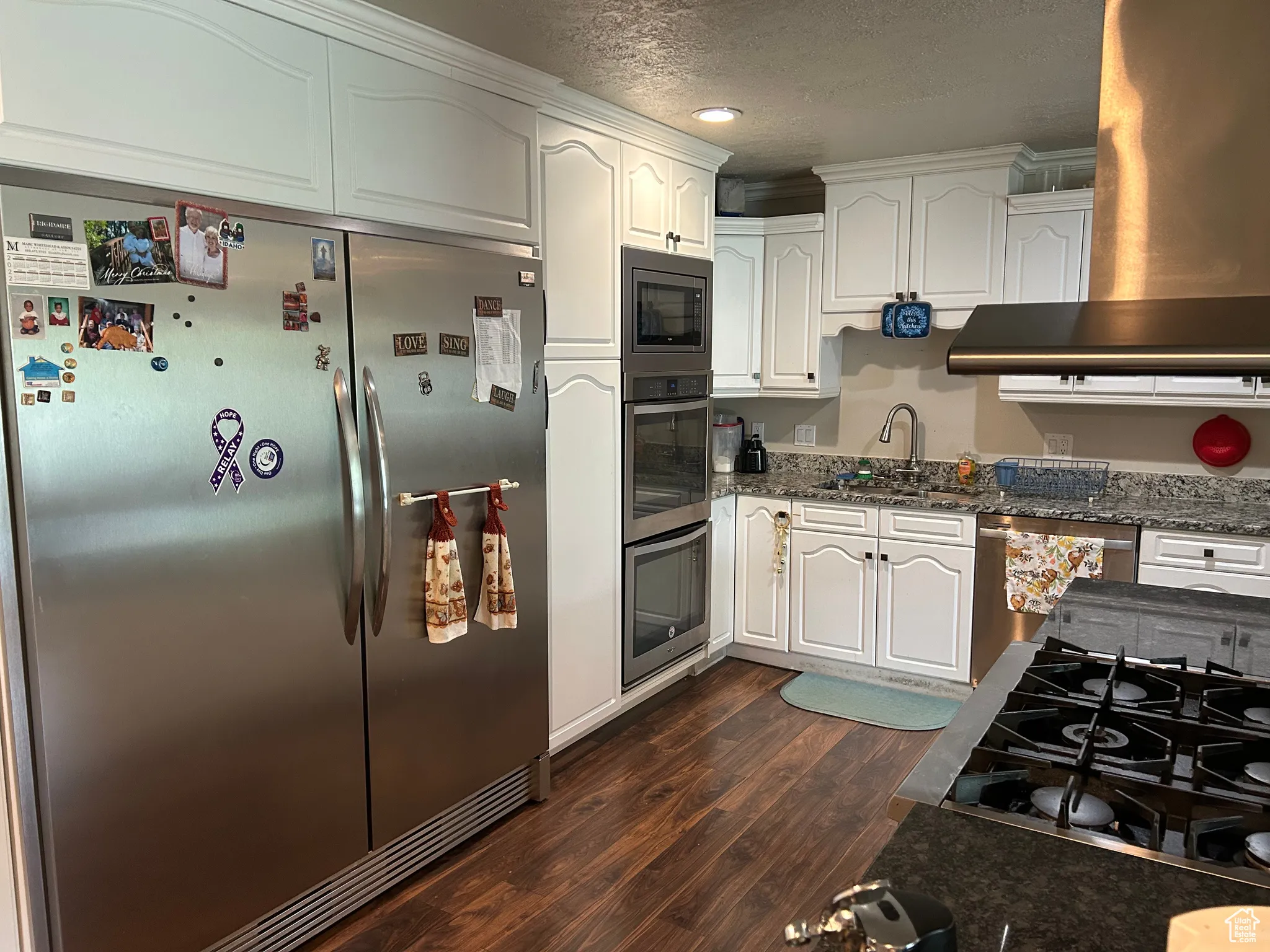 Kitchen with appliances with stainless steel finishes, dark wood-style flooring, a sink, white cabinets, and wall chimney range hood