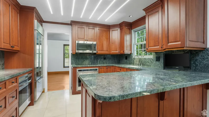 Kitchen with appliances with stainless steel finishes, brown cabinets, decorative backsplash, a peninsula, and light tile patterned flooring