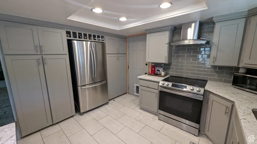 Kitchen featuring stainless steel appliances, gray cabinetry, wall chimney exhaust hood, a tray ceiling, and recessed lighting