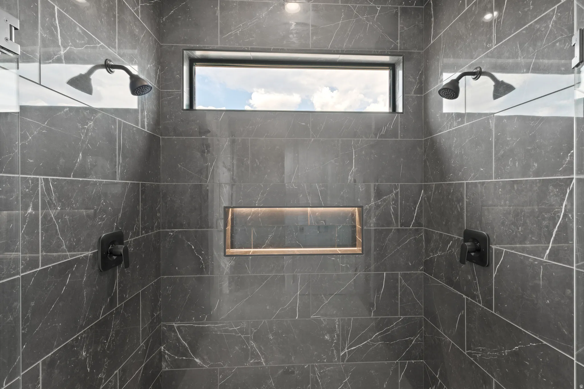 Dual matte black shower heads, dark marble-look tile, built-in shower niche with accent lighting, and transom window