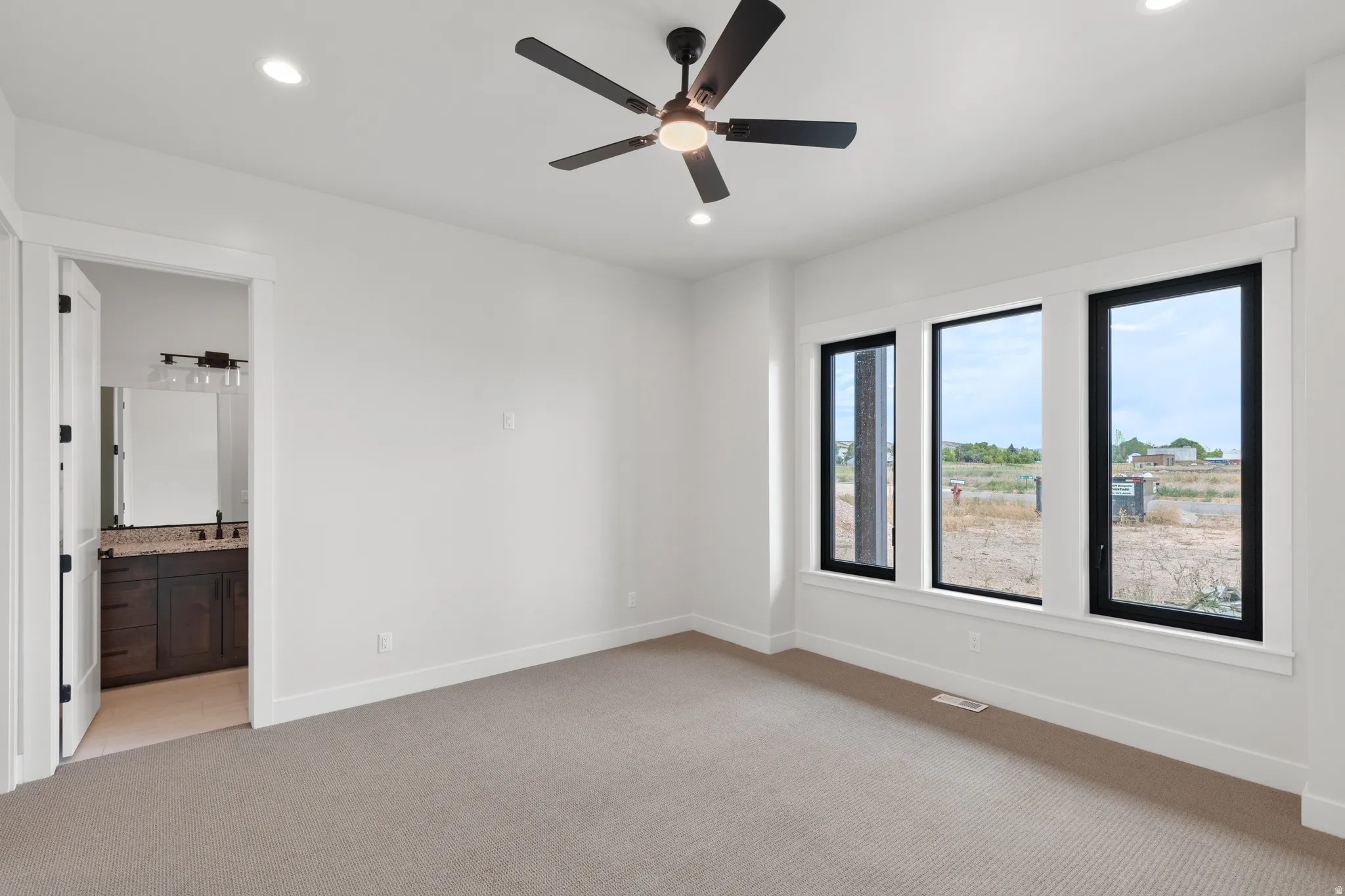Recessed lighting, ceiling fan with integrated light, three large windows with black frames, carpeted flooring, and a doorway revealing a vanity with a dark wood-finish cabinet and stone countertop