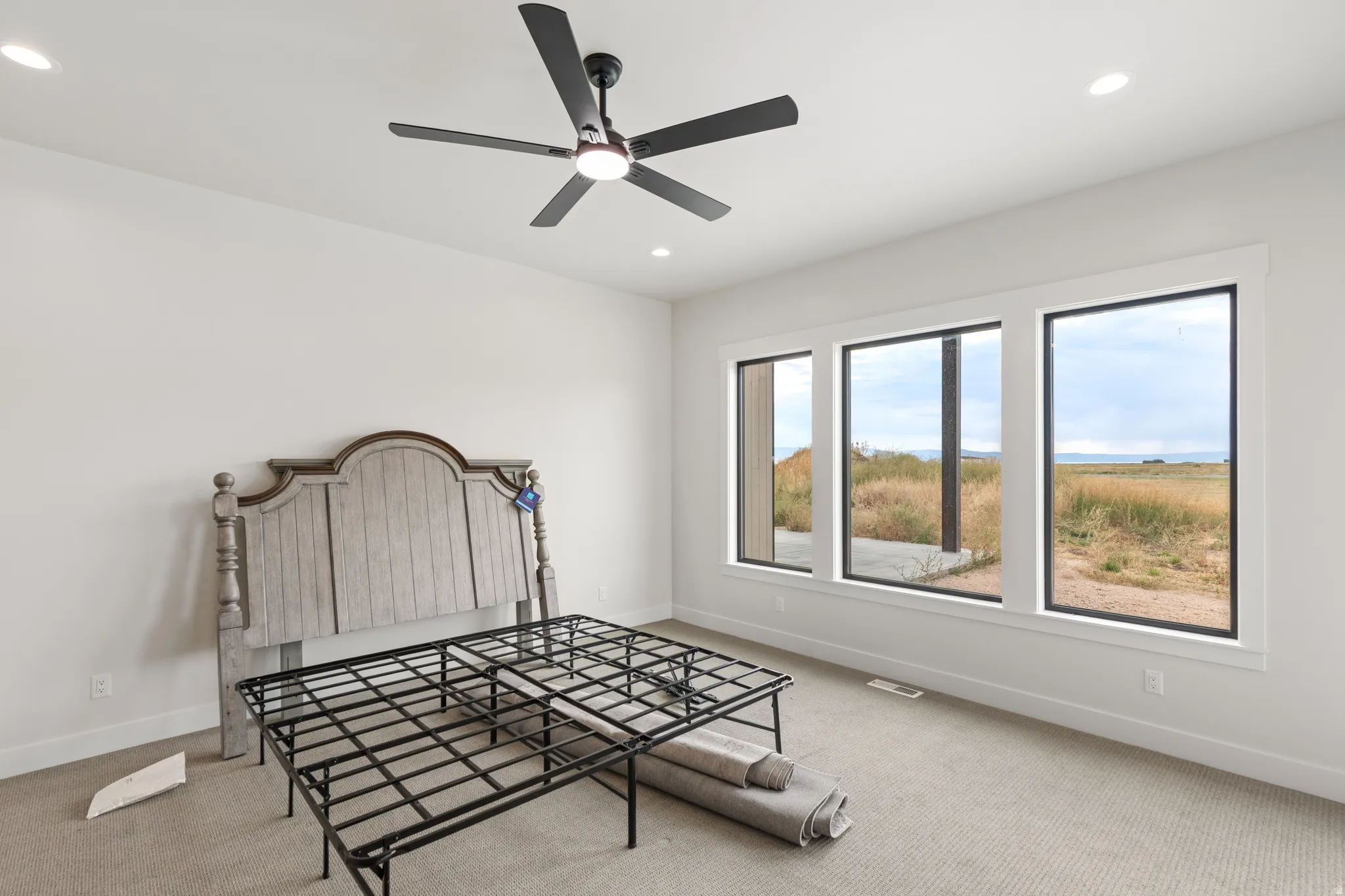 Recessed ceiling lighting, ceiling fan with integrated light, three large windows with black framing, light neutral-toned carpet flooring, and white painted walls