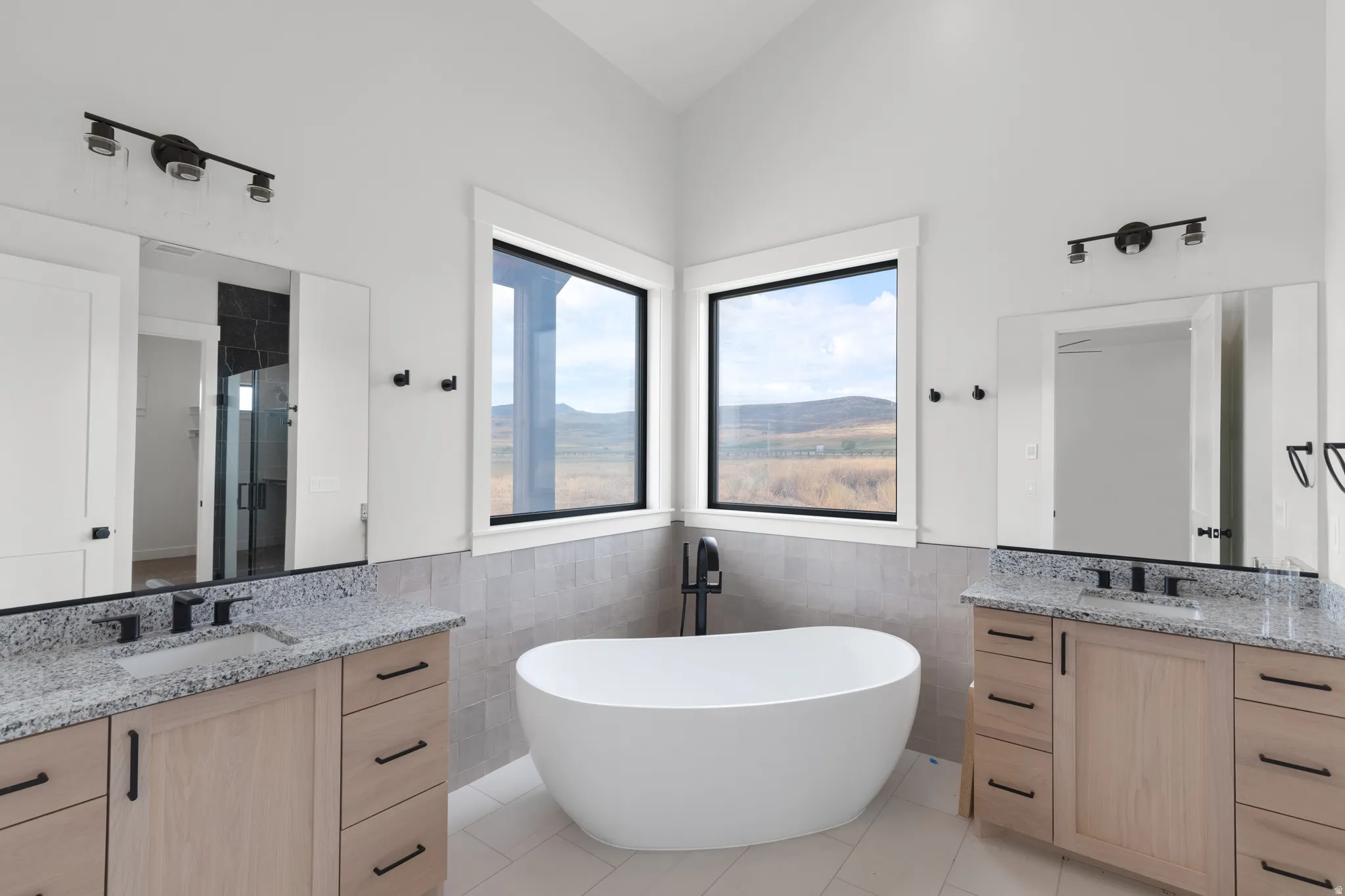 Dual light wood-finish vanities with granite countertops, a freestanding soaking tub, and large corner windows