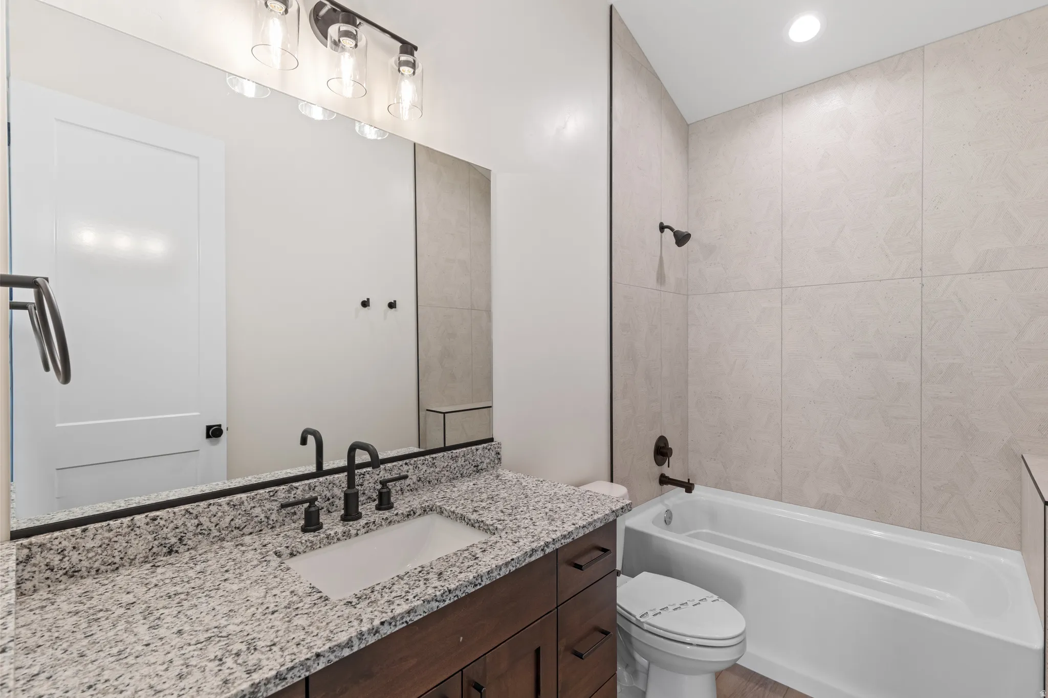 White rectangular undermount sink with a black faucet, granite-style countertop, dark wood-finish vanity with drawer storage, large wall mirror, and a white bathtub with a black shower head