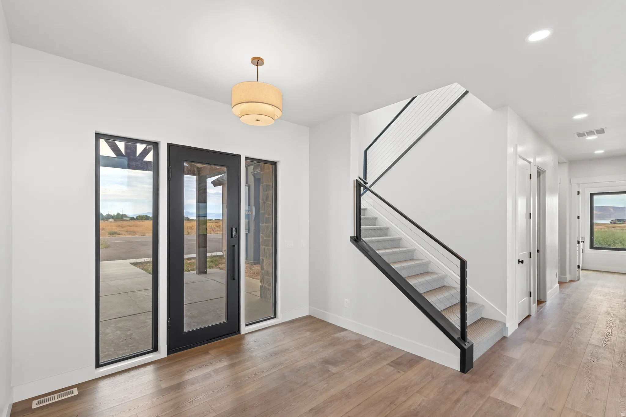 Wood-finish flooring throughout, modern staircase with black railing, recessed lighting, contemporary front door with sidelights, and a tiered semi-flush mount light fixture